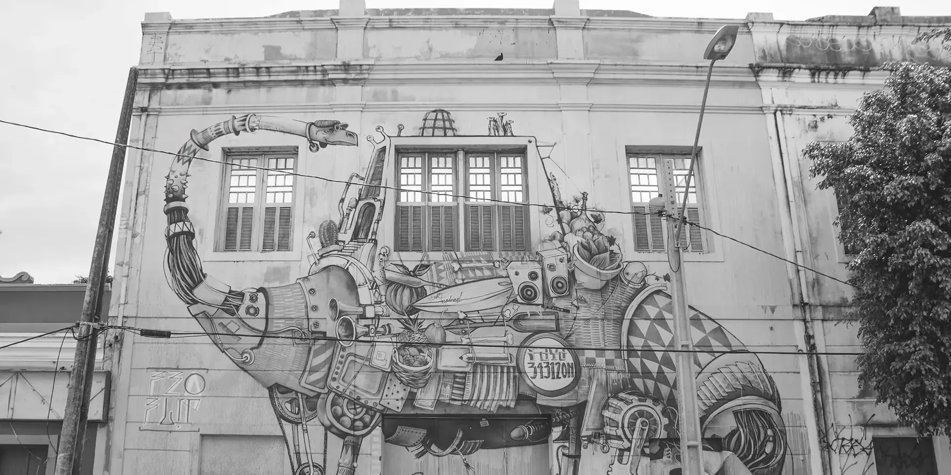 Fotografia em tons de cinza de um grande mural na fachada de um prédio antigo com um dinossauro futurista com vários acessórios sendo observado por um grupo de pessoas na frente do prédio