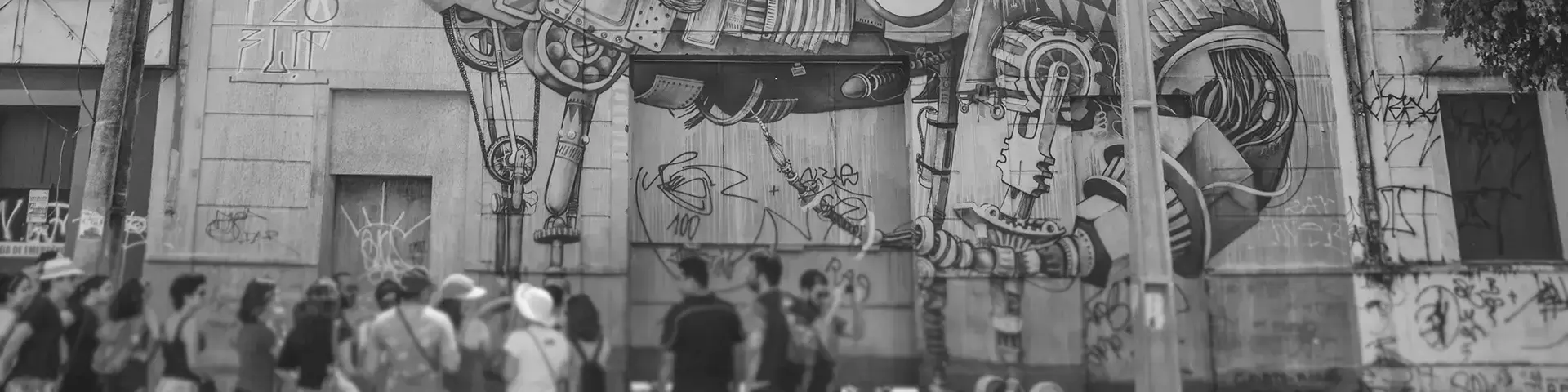 Fotografia em tons de cinza de um grande mural na fachada de um prédio antigo com um dinossauro futurista com vários acessórios sendo observado por um grupo de pessoas na frente do prédio