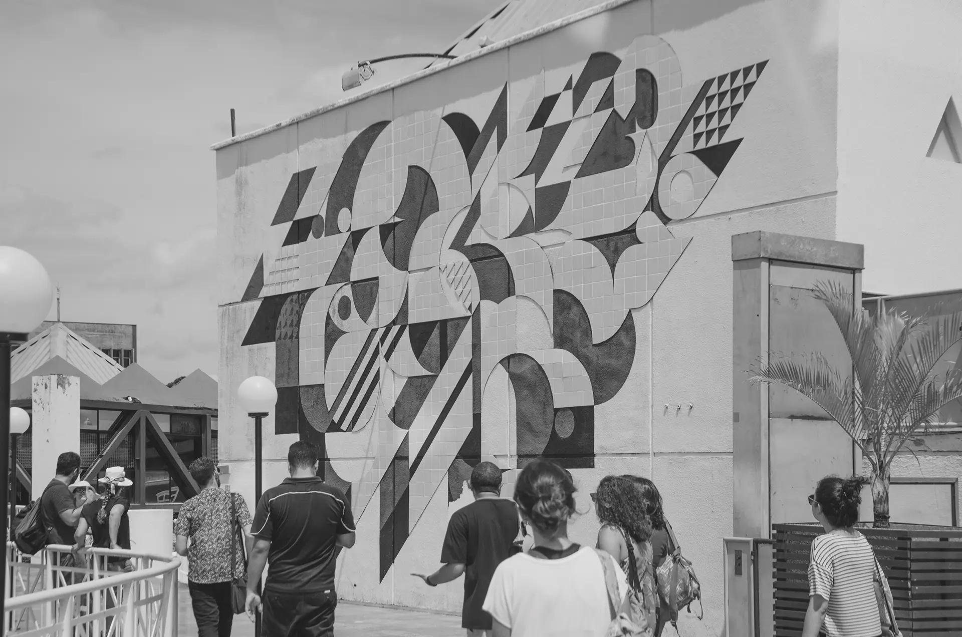 Fotografia em tons de cinza de um mural com pinturas geométricas e azulejos com um grupo de pessoas de costas observando o mural