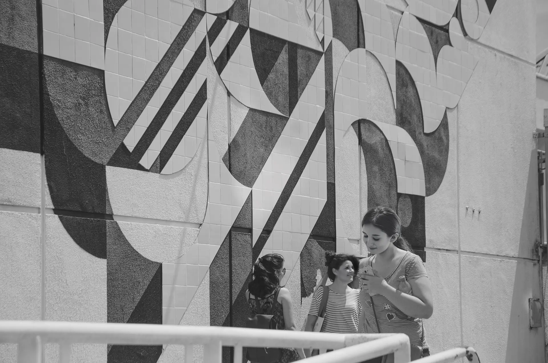Fotografia em tons de cinza de um mural com pinturas geométricas e azulejos com um grupo de pessoas de costas observando o mural