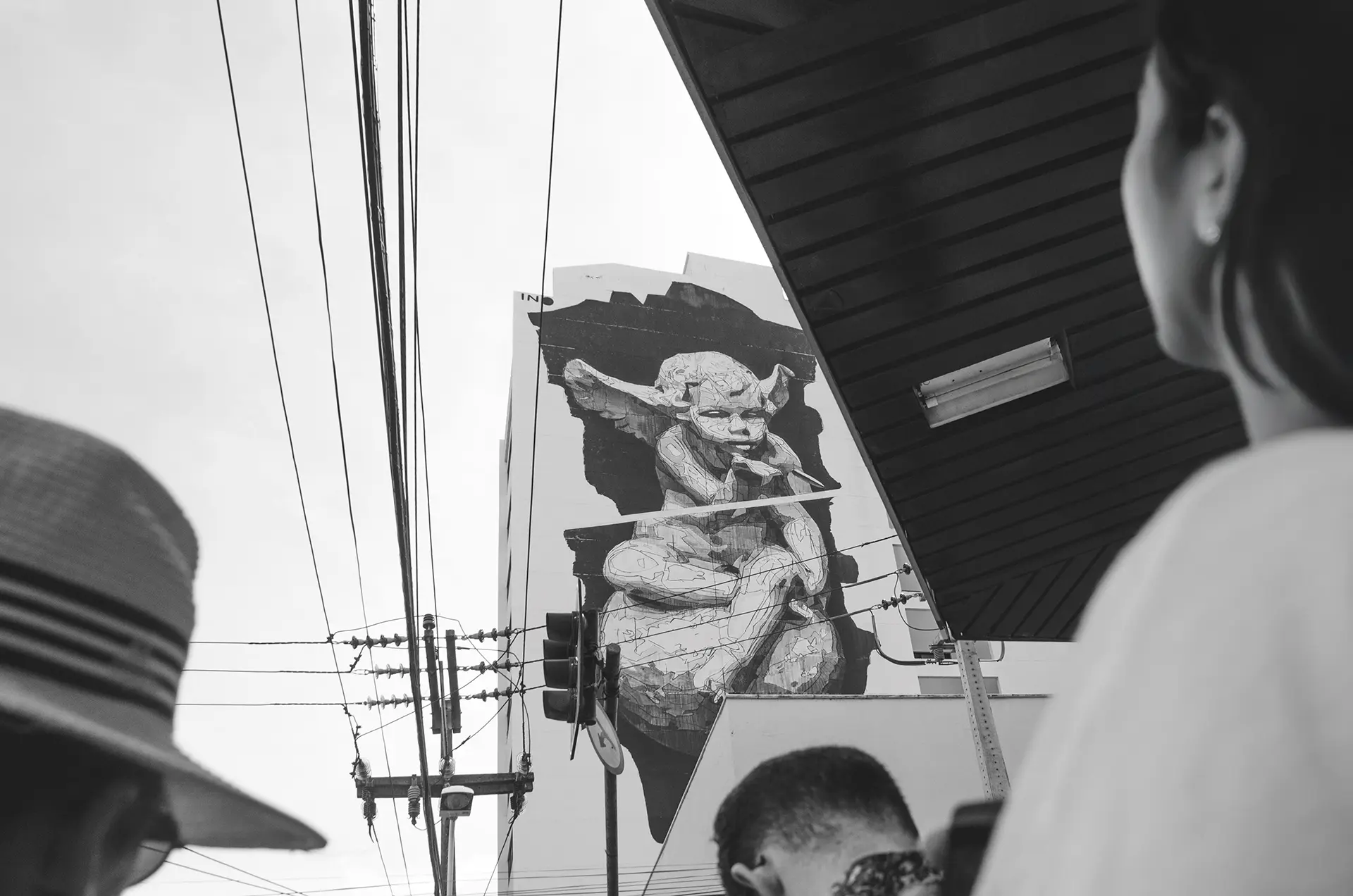 Fotografia em tons de cinza de um gigante mural na lateral de um prédio com um anjinho assoprando uma bala no Centro de Fortaleza com um homem de costas com a cabeça baixa