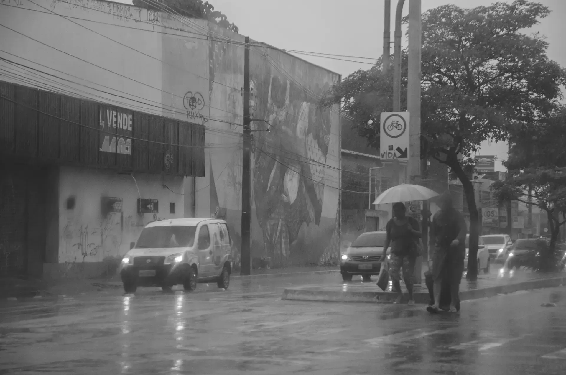 Fotografia em tons de cinza de um cruzamento do bairro Benfica em Fortaleza onde é possível observar carros, pedestres e um grafitti na parede de um comércio ao fundo