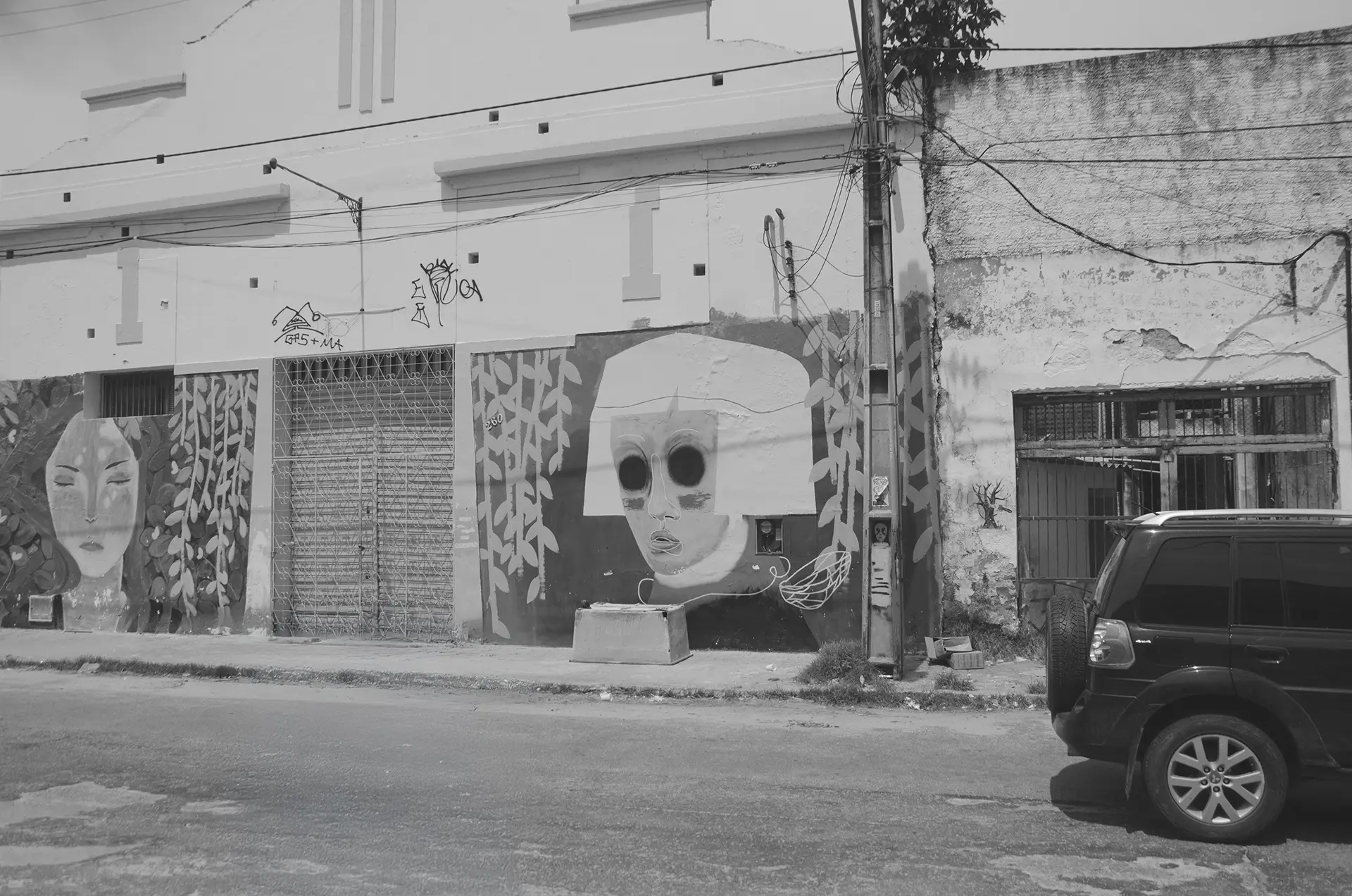 Frame 1 em tons de cinza de uma sequencia que mostra um carro atravessando uma rua no Centro de Fortaleza da esquerda para a direita com um mural de graffiti ao fundo