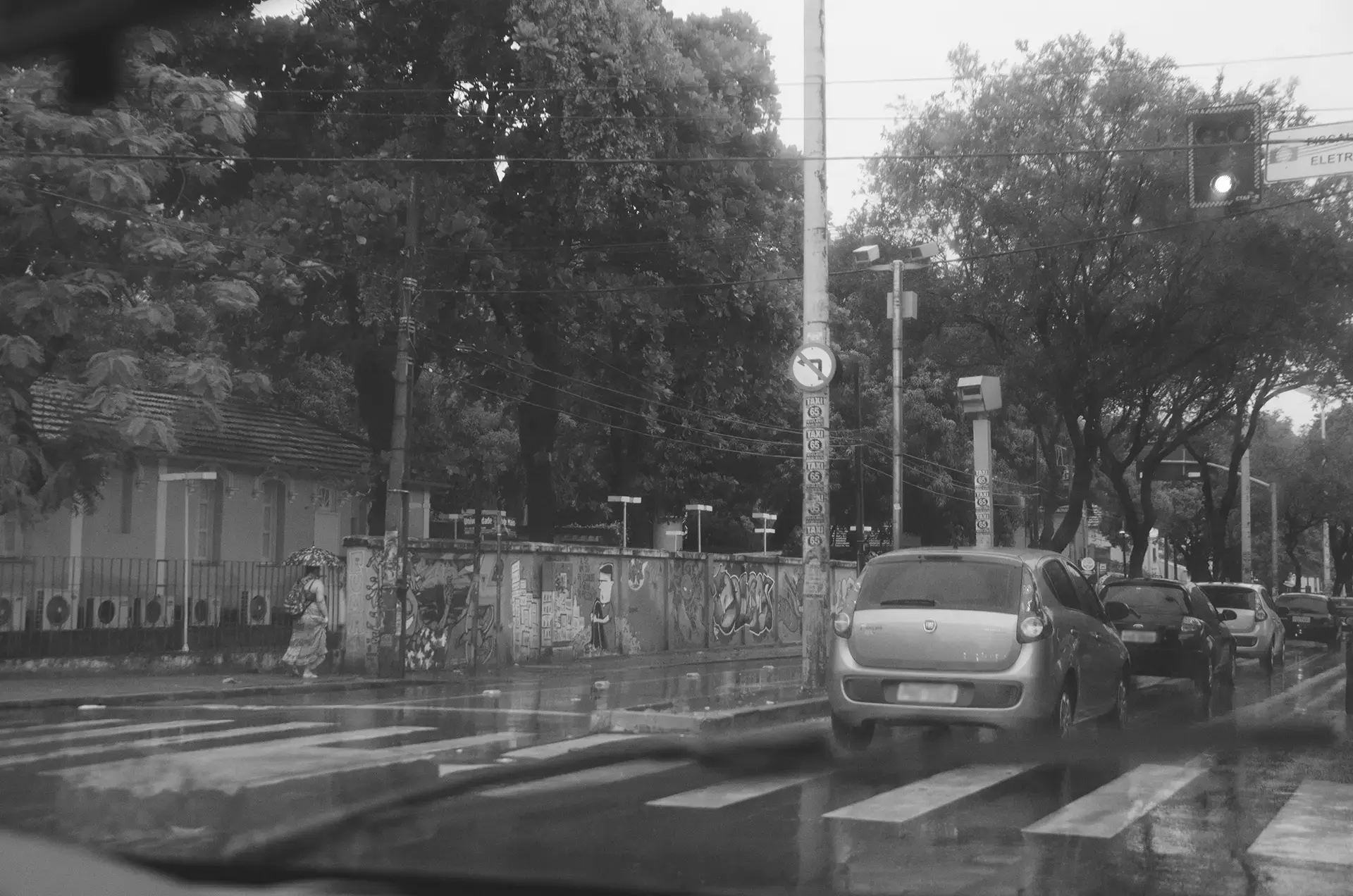 Fotografia em tons de cinza de carros parados em um sinal na Avenida 13 de Maio em Fortaleza durante uma tarde chuvosa onde é possível observar uma pedestre com seu guarda chuva e os muros da Universidade Federal do Ceará com intervenções de arte urbana