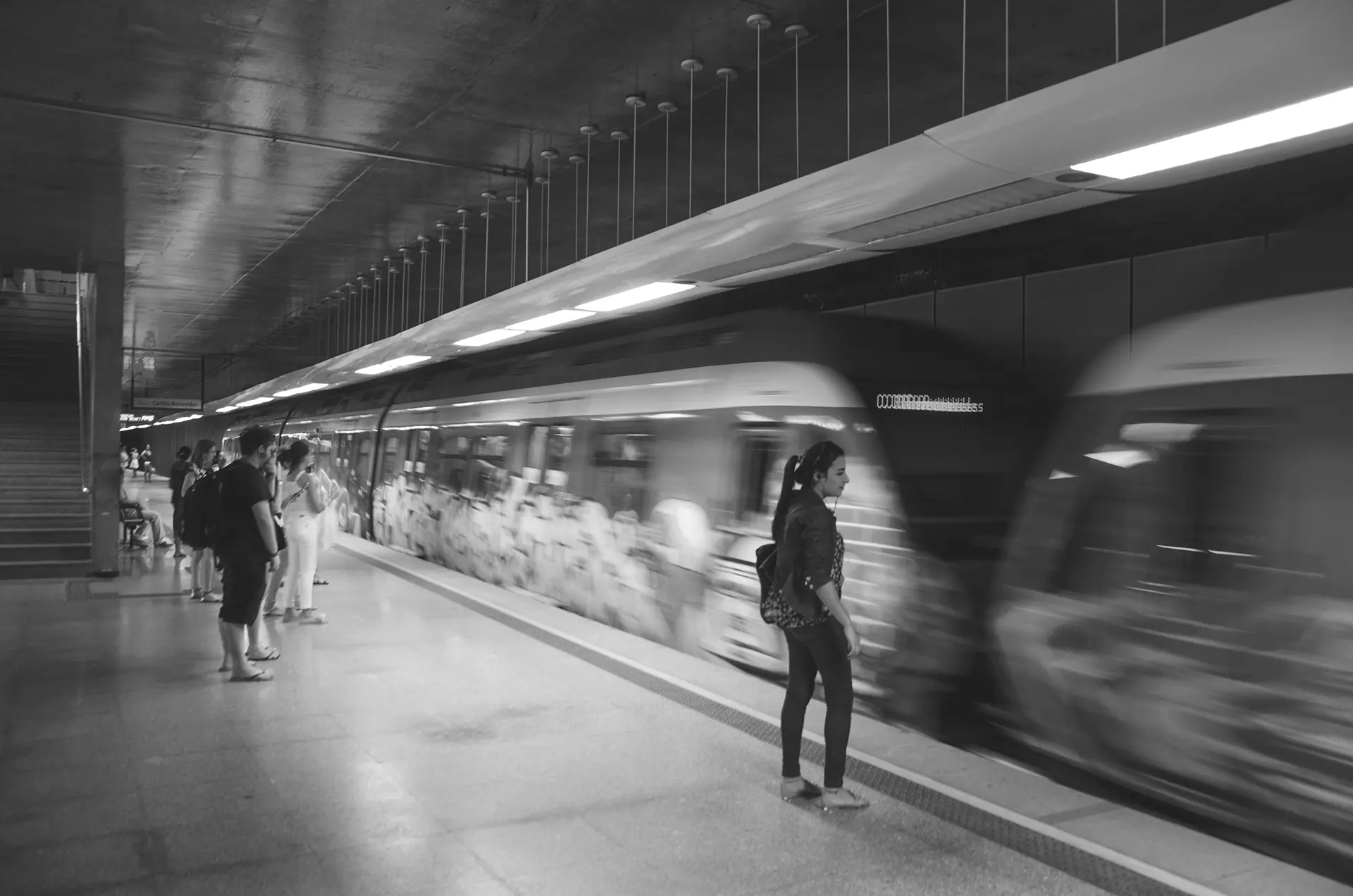Fotografia em tons de cinza de uma mulher esperando o trem para na Estação de metrô do Benfica em Fortaleza onde é possível observar os vagões grafitados do trem