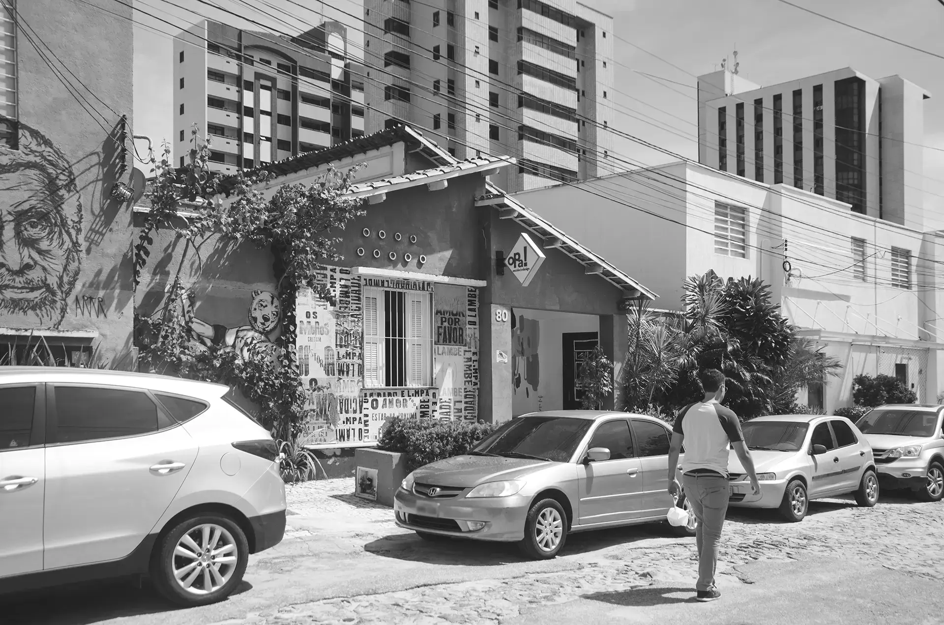 Fotografia em tons de cinza de uma viela onde é possível observar carros estacionados, um transeunte passando e os muros com intervenções de arte urbana
