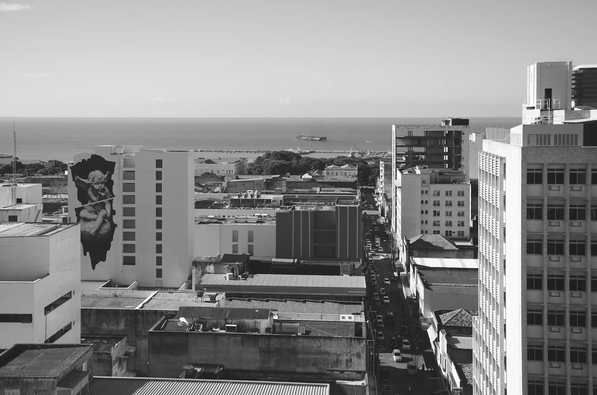 Fotografia em tons de cinza em perspectival do Centro de Fortaleza onde é possível observar vários prédios e um deles com um mural de uma anjinho assoprando uma bala e a orla da cidade ao fundo