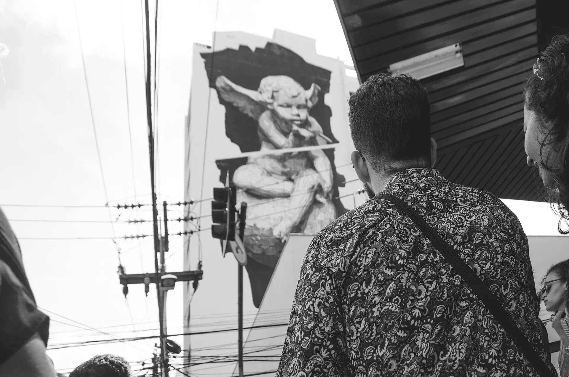 Fotografia em tons de cinza de um gigante mural na lateral de um prédio com um anjinho assoprando uma bala no Centro de Fortaleza com um homem de costas o observando