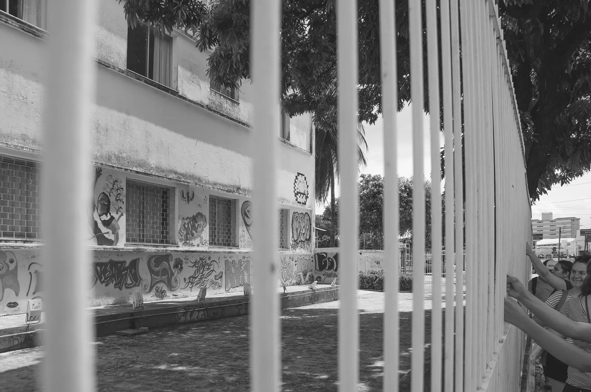 Fotografia em tons de cinza em perspectiva de uma grade com pessoas segurando-a e ao fundo um prédio com intervenções de arte urbana