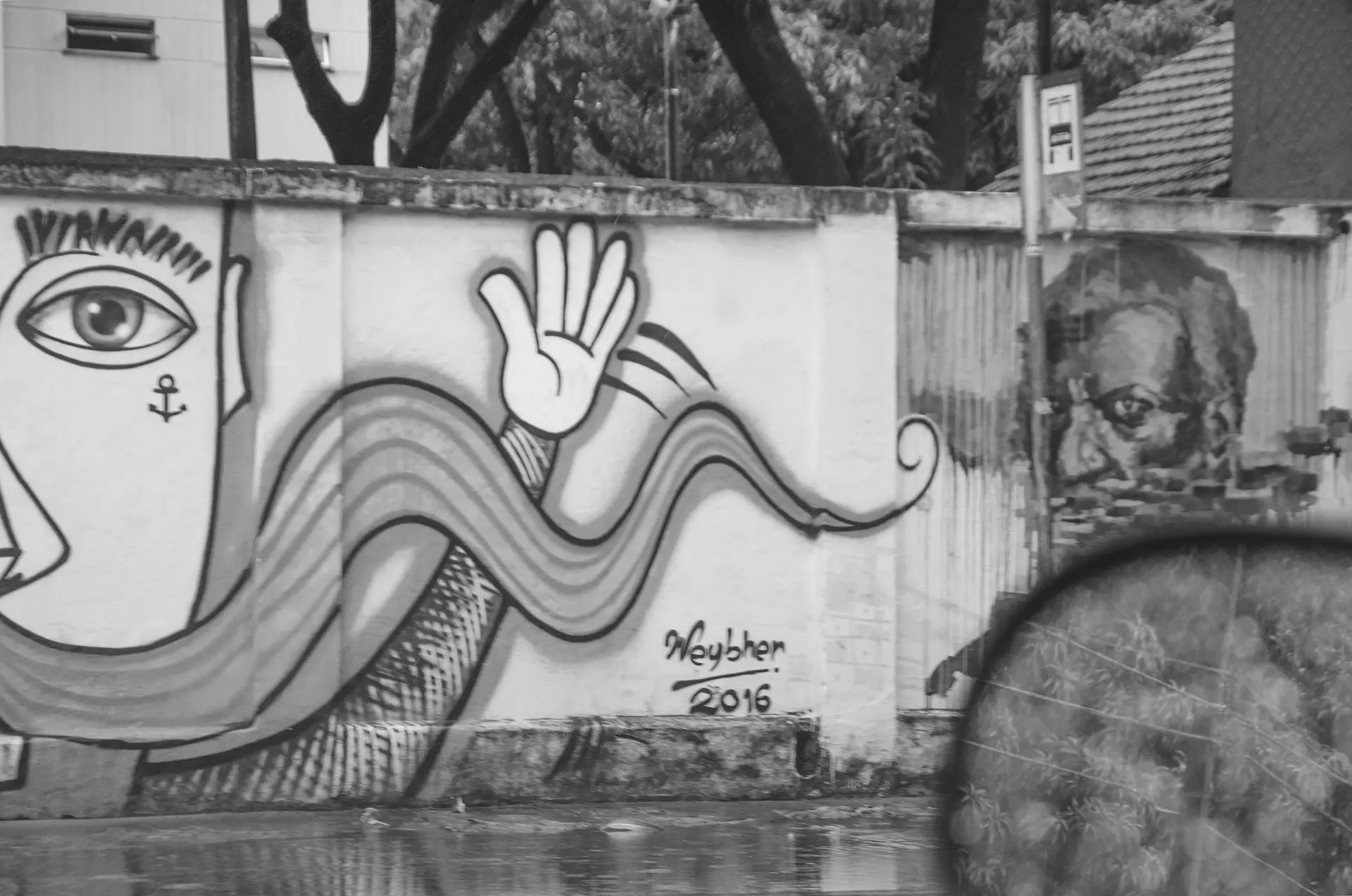 Fotografia em tons de cinza de um muro grafitado na Avenida 13 de Maio em Fortaleza feito através da janela de um carro onde é possível observar o retrovisor e ao fundo um dos murais de um homem preto olhando atentamente de volta
