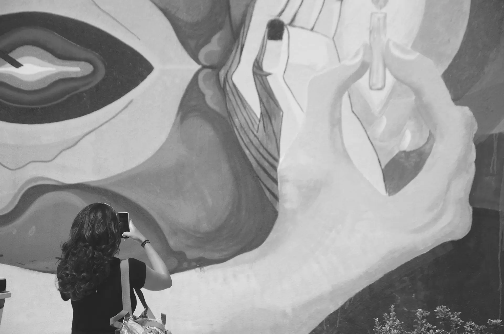 Fotografia em tons de cinza de uma mulher de costas fotografando um mural de graffiti no muro de uma praça no Centro de Fortaleza com o celular