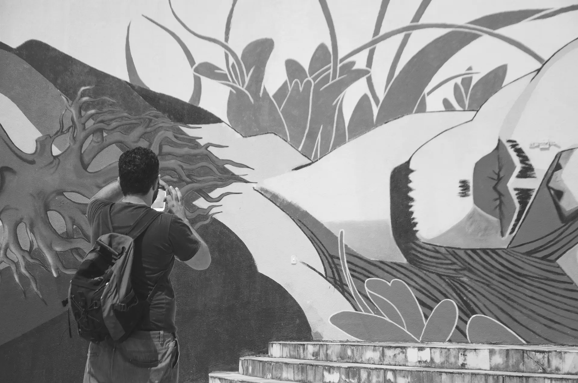 Fotografia em tons de cinza de um homem de costas fotografando um mural de graffiti no muro de uma praça no Centro de Fortaleza com o celular