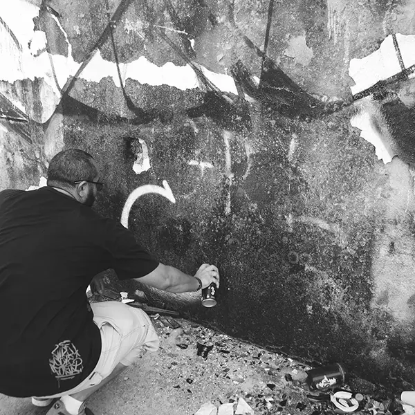 Frame 1 em tons de cinza de um vídeo de um homem de cócoras com uma lata de spray na mão enquanto desenha uma assinatura em um muro de um prédio abandonado