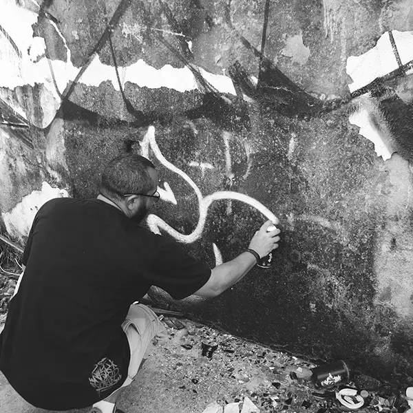 Frame 10 em tons de cinza de um vídeo de um homem de cócoras com uma lata de spray na mão enquanto desenha uma assinatura em um muro de um prédio abandonado