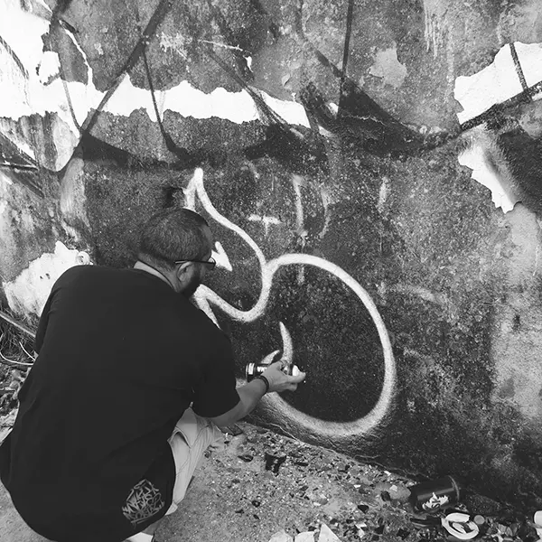 Frame 13 em tons de cinza de um vídeo de um homem de cócoras com uma lata de spray na mão enquanto desenha uma assinatura em um muro de um prédio abandonado