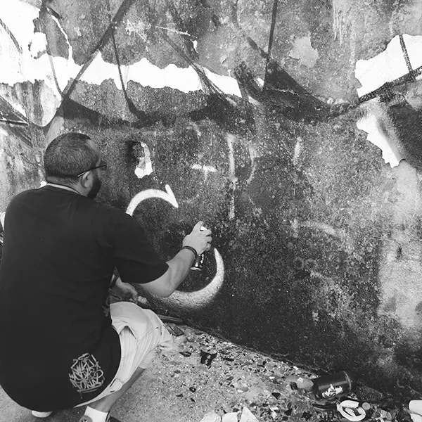 Frame 4 em tons de cinza de um vídeo de um homem de cócoras com uma lata de spray na mão enquanto desenha uma assinatura em um muro de um prédio abandonado