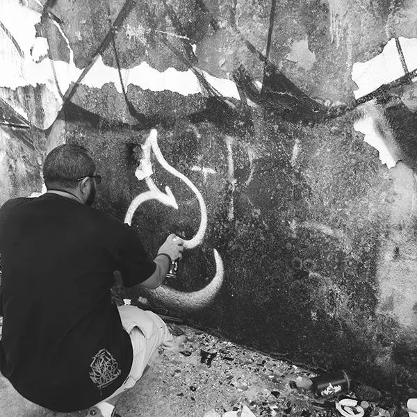Frame 7 em tons de cinza de um vídeo de um homem de cócoras com uma lata de spray na mão enquanto desenha uma assinatura em um muro de um prédio abandonado