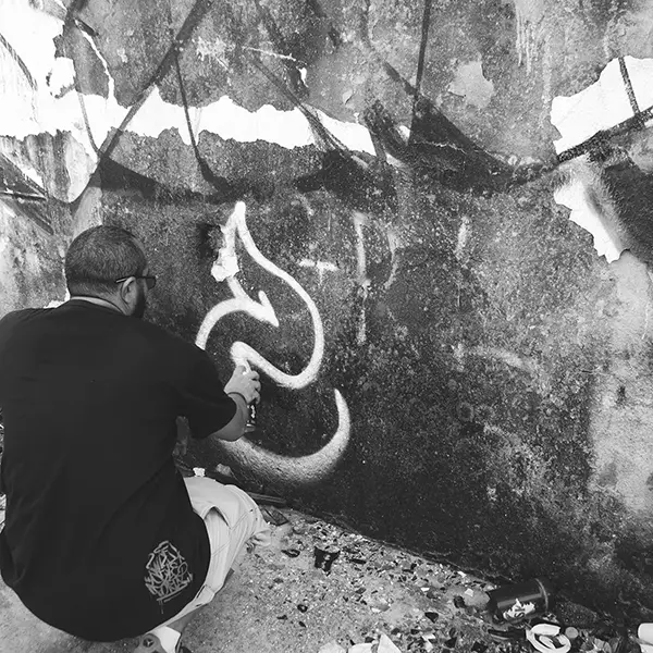 Frame 8 em tons de cinza de um vídeo de um homem de cócoras com uma lata de spray na mão enquanto desenha uma assinatura em um muro de um prédio abandonado